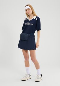 Kobieta w granatowej sukience z logo 'ellesse', białą opaską na głowę, wysokimi skarpetkami, białymi sportowymi butami i granatową saszetką na talię, stojąca na jednolitym tle.