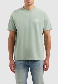 Lichtgroene katoenen T-shirt met korte mouwen, ronde hals en een klein wit logo op de linkerborst. Gecombineerd met vervaagde blauwe jeans.