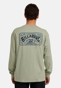 Uomo che indossa una camicia a maniche lunghe verde chiaro con una grafica sul retro raffigurante un tramonto, onde, palme e la scritta "Billabong".