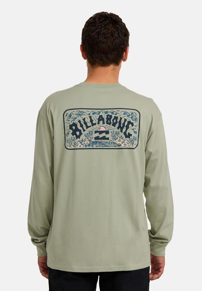 Mann trägt ein hellgrünes Langarmshirt mit einem Rückenmotiv von einem Sonnenuntergang, Wellen, Palmen und dem Wort "Billabong."