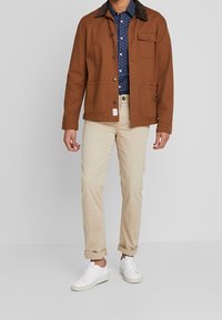 Hnědá plátěná bunda s tmavě hnědým límcem, dvěma předními kapsami, v kombinaci s tmavě modrou košilí s puntíky, béžovými kalhotami a bílými teniskami.