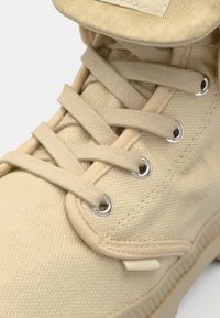 Baskets montantes en tissu beige avec bout arrondi, œillets métalliques, lacets plats et languette rembourrée avec une étiquette. Surfaces texturées sur l'ensemble.