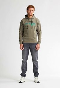 Olivgrön hoodie med dragsnören och tryckt text, kombinerad med mörkgrå jeans och vita sneakers. Avslappnad, ledig passform.