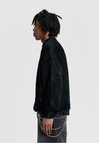 Schwarzer strukturierten Pullover mit lockerer Passform, runder Ausschnitt und gerippten Bündchen. Kombiniert mit dunklen Jeans und einem silbernen Kettenaccessoire.