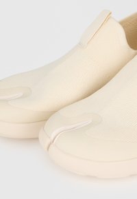 Κοντινή λήψη μπεζ παπουτσιών τύπου slip-on με υφασμάτινο επάνω μέρος με υφή και σχεδιασμό διαχωρισμένου δαχτύλου σε ανοιχτό φόντο.