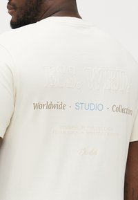 Krem bomulls t-skjorte med brodert tekst i ulike farger på baksiden, som fremhever merkevarens detaljer og designopprinnelse.