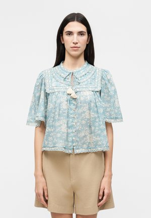 Femme aux cheveux raides et foncés portant un chemisier bleu clair à motifs floraux avec des pompons et un short beige, debout devant un fond uni.