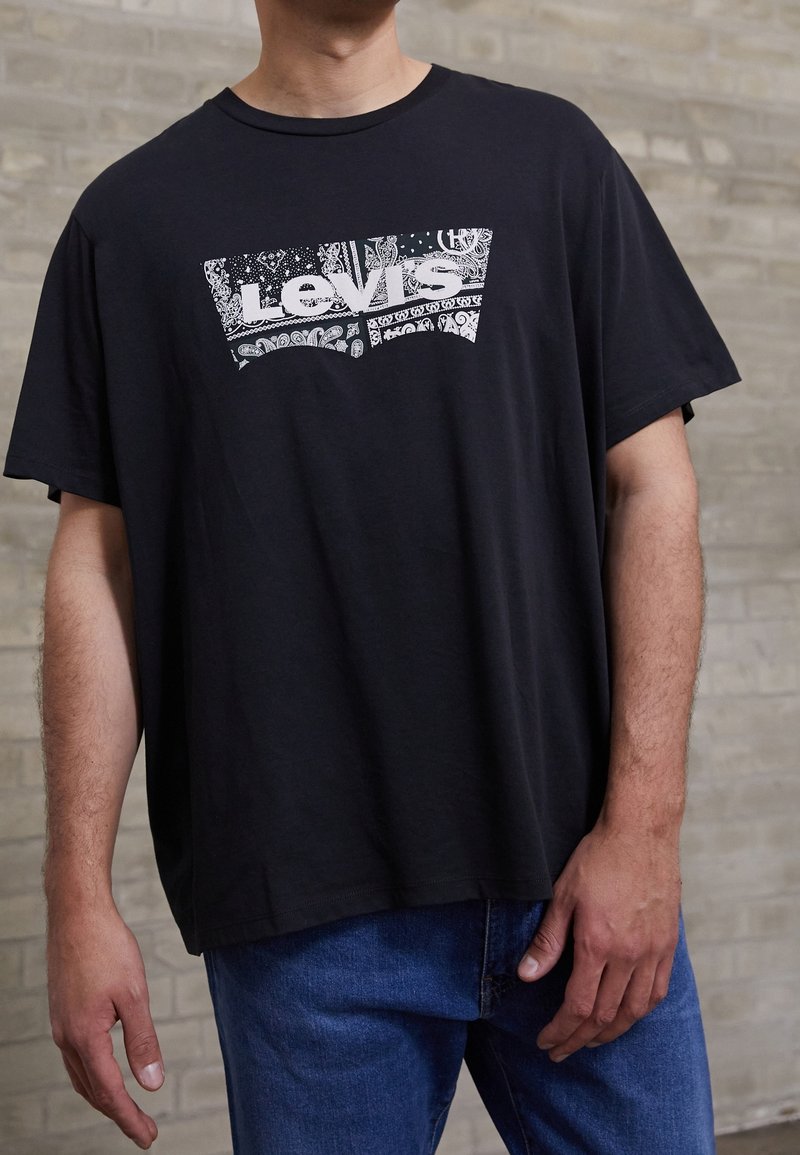 Homme portant un T-shirt noir Levi's avec un logo paisley blanc et un jean bleu, se tenant devant un mur en briques claires.