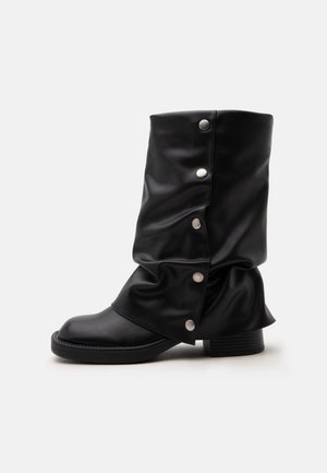 Botte en cuir noire jusqu'à mi-mollet, ample, avec boutons-pression argentés le long du côté extérieur et petit talon empilé sur un fond uni.