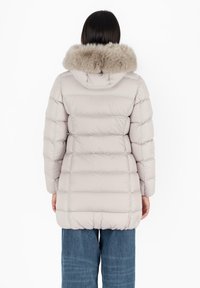 Giacca puffer grigio chiaro con cappuccio bordato di pelliccia, design squadrato, texture trapuntata e orlo elastico. Indossata con pantaloni di denim blu.