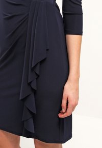 Tummansininen mekko, jossa on kolmenneljäsosakäsihihat, ja siinä on pystysuora röyhelökohta hameen alaosassa rentona käden lähellä.