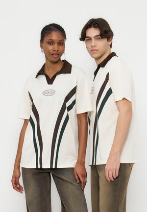 To modeller står side om side iført matchende hvide kortærmede skjorter med brune kraver og lodrette mørke striber.