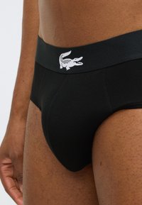 Zwarte slip van een gladde stof, met een contrasterend wit krokodillenlogo op de tailleband. Traditionele, aangesloten pasvorm met onopvallende naden.