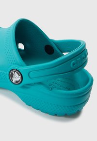 Türkiissinised Crocsid ümarate servadega, tekstuurse pinnaga ja taga oleva rihmaga. Omavad musta nuppu logoga. Materjal on paindlik ja kerge.