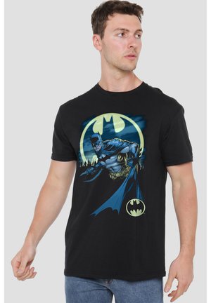 Mann trägt schwarzes T-Shirt mit Batman-Grafik und Logo, schaut nach rechts, steht vor einfachem hellem Hintergrund.