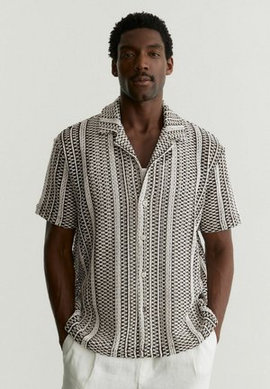 Muž v krátkym rukávom čierno-bielom vzorovanom košeli na gombíky a bielych nohaviciach, stojaci s rukami v vreckách na jednoliatom pozadí.