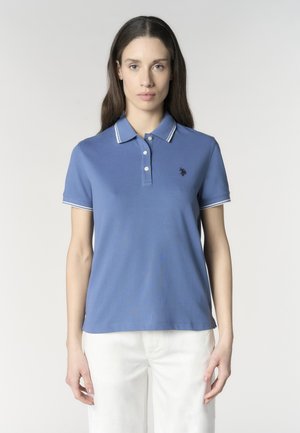 Donna con lunghi capelli castani che indossa una polo blu a maniche corte con bordi bianchi e pantaloni bianchi, in piedi davanti a uno sfondo semplice.