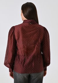 Bordeauxrote Bluse mit Puffärmeln, transparenter Stoff, gepunktetes Muster, hoher Kragen und Knopfdetails auf der Rückseite, kombiniert mit grauen Jeans.