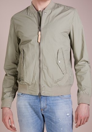 Personne portant une veste bomber marron clair avec des poches frontales et un jean bleu clair usé.