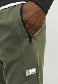 Olijfgroene joggers met ritszak aan de zijkant, gestructureerde afwerking en een wit logo-label op de linkerdij.
