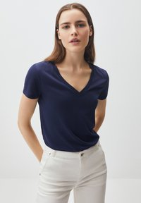 T-shirt a V con collo navy realizzata in tessuto liscio, maniche corte, forma aderente. Indossata con pantaloni dritti di colore chiaro. Design semplice, dettagli minimi.