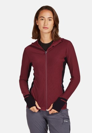 Giacca a zip con cappuccio color ruggine, in tessuto testurizzato, con dettagli neri, polsini con foro per il pollice e tasche frontali, abbinata a pantaloni grigi.