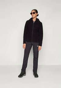 Svart fleecejakke med glidelås foran og hette, kombinert med mørke bukser og svart fottøy. Modell med solbriller. Elegant, moderne design.