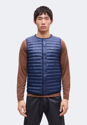 Homme portant une veste matelassée sans manches bleue par-dessus une chemise marron à manches longues avec un pantalon noir, debout devant un fond uni.