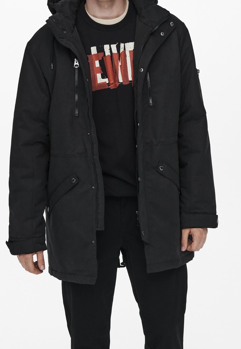 Homme portant une parka noire à capuche sur un t-shirt graphique noir et un pantalon noir, sur fond blanc.