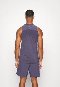 Man som står med ryggen mot kameran, iklädd en mörk lila atletisk linne och matchande shorts, mot en enkel vit bakgrund.