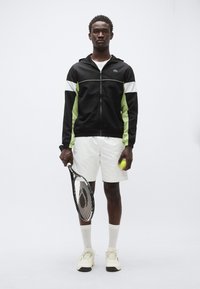 Črno-zelen jopič z zadrgo, bele kratke hlače, drži teniški lopar in žogico. Ponaša se z teksturiranim tkanino, športnim dizajnom in kontrastnimi poudarki.