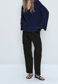 Marineblaue gerippte Strickjacke mit V-Ausschnitt, kombiniert mit schwarzen Cargo-Hosen mit seitlichen Taschen und flachen schwarzen Sandalen.