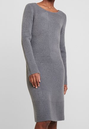 Nainen, joka pukeutunut pitkähihainen, polvipituinen harmaa neulemekko, jossa on resorit hihoissa ja helmassa, seisoo yksinkertaisen taustan edessä.