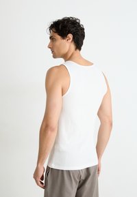 Weißes Baumwoll-Top mit gerippter Textur, rundem Ausschnitt und breiten Trägern, getragen mit grauen Hosen. Körper ist von der Kamera abgewandt.