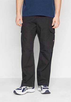 Mann in schwarzen Cargo-Hosen und weißen, marineblauen sowie grünen Laufschuhen, der vor einem schlichten Hintergrund steht.