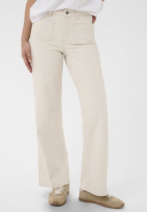 Person iført beige bukser med høj talje og lige ben samt beige sneakers, stående foran en ensfarvet hvid baggrund.