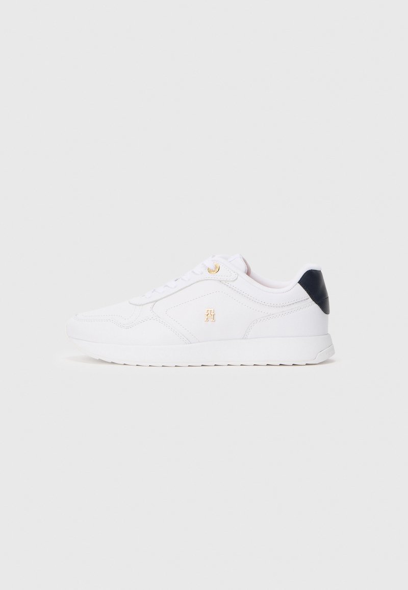 Λευκό χαμηλό sneaker με χρυσά μάτια και λεπτομέρειες λογότυπου, ναυτικό μπλε πίσω τμήμα και λευκή σόλα, εμφανιζόμενο από την εξωτερική πλευρά σε ουδέτερο φόντο.