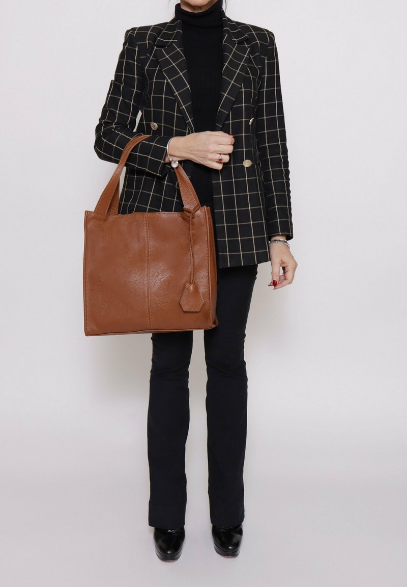 Bruine leren tote bag met een gladde textuur, vierkante vorm en twee stevige handvatten. Heeft een decoratief label en minimale hardwareaccenten.