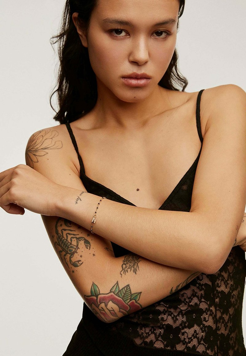 Camisón de encaje negro con tirantes finos; los brazos del modelo adornados con tatuajes coloridos y una delicada pulsera en una muñeca.