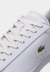 Baskets blanches Lacoste avec lacets blancs, nom de marque embossé sur la languette et logo crocodile vert sur le côté.