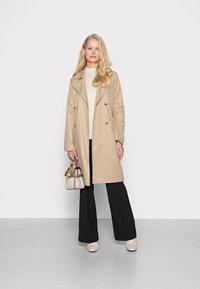 Trench coat bej cu două rânduri de nasturi, croială ajustată, cu guler. Asortat cu un pulover ușor, pantaloni negri cu picioare largi și o mică geantă bej.