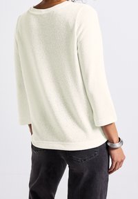 Pull texturé de couleur crème avec manches trois-quarts, encolure arrondie et coupe décontractée, associé à un jean foncé. Bracelet en argent visible.