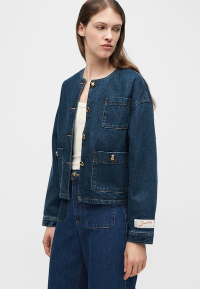 Γυναίκα που φοράει σκούρο μπλε τζιν μπουφάν με μεγάλες τσέπες και χρυσά κουμπιά, συνδυασμένο με τζιν παντελόνι στο ίδιο χρώμα και λευκό τοπ από μέσα.