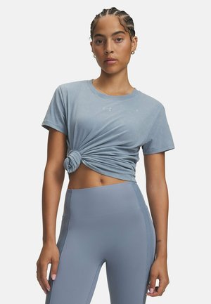 Vrouw met gevlochten haar, die een lichtblauw geknoopt T-shirt en hoge taille blauwe leggings draagt, staande tegen een effen achtergrond.