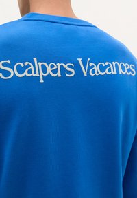 Sweatshirt albastru royal cu text alb "Scalpers Vacances" pe spate, confecționat dintr-un material neted și texturat, cu un design tip crew neck.