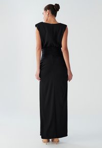 Abito da sera nero con silhouette aderente, maniche a cappuccio e un design elegante sul retro. Il tessuto è liscio con una leggera lucentezza.