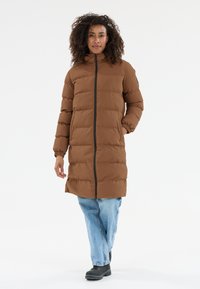 Kvindelig med krøllet hår iført en brun lang pufferjakke, lyseblå jeans og sorte støvler, stående med den ene hånd i lommen.