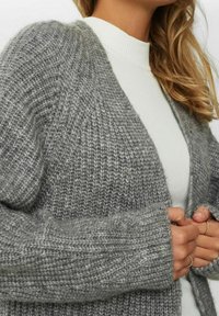 Ljusgrå stickad cardigan med en strukturerad mönstring, tjocka långa ärmar och ribbade ärmslut. Bärs över en vit topp med hög hals.