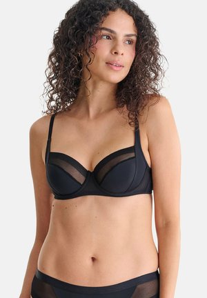 Femme aux cheveux bouclés portant un soutien-gorge en maille noire et tissu uni ainsi qu'une culotte taille haute assortie, regardant légèrement sur le côté.