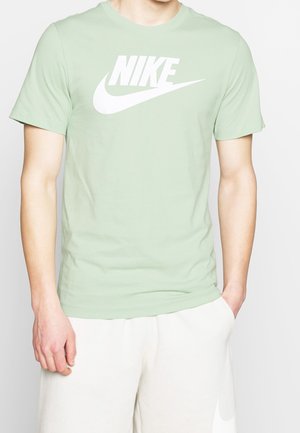 Personne portant un t-shirt Nike vert clair avec logo blanc et un short blanc, une main dans la poche, devant un fond uni.